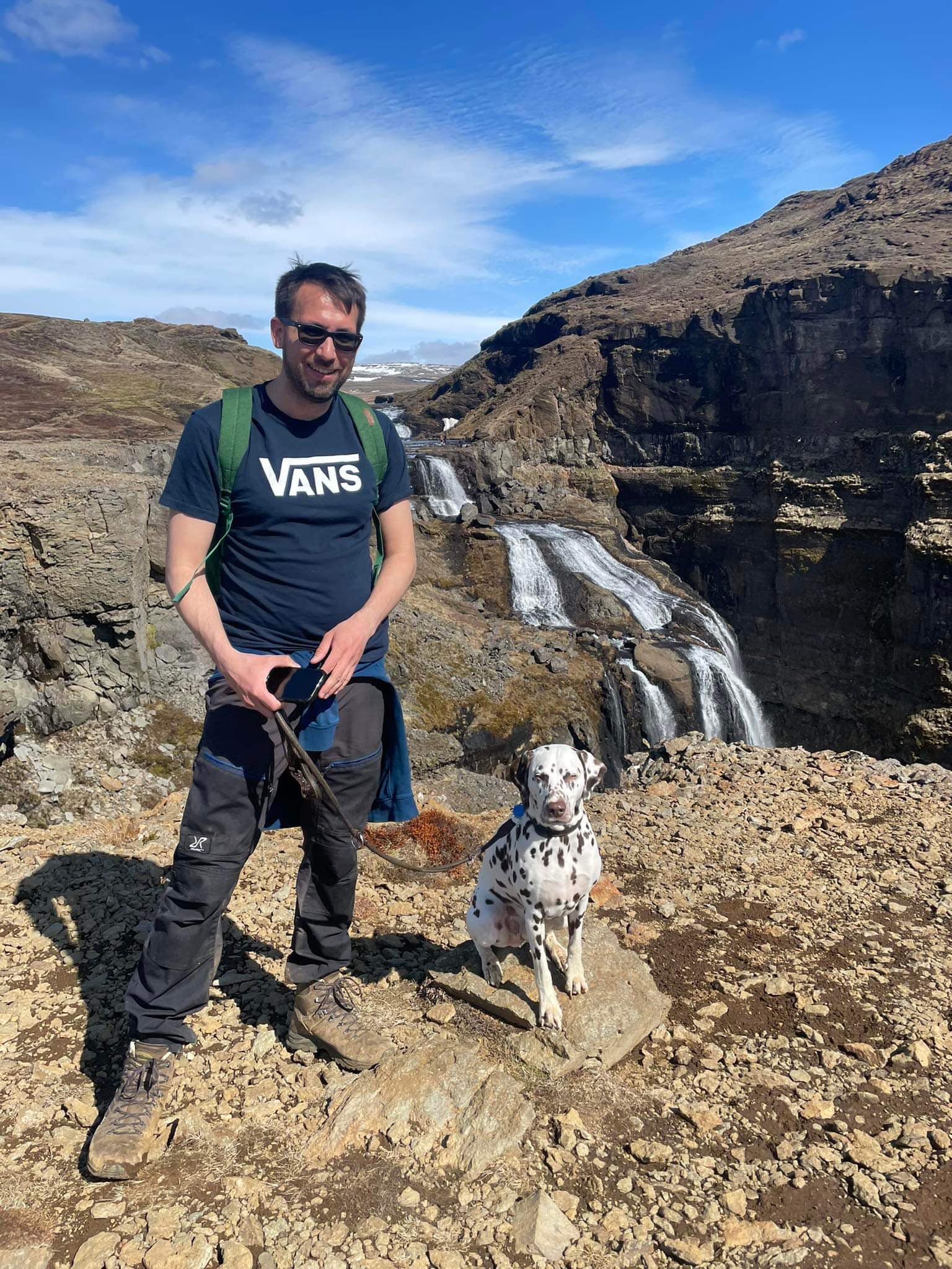 Man in blue shirt with a brown spotted dalmatian on the leash next to him, a mountain range in the back with a waterfall